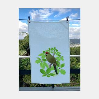Pale blue tea towel featuring an illustration of a kākāriki perched amongst whauwhaupaku leaves, displayed on clothesline overlooking the Hutt Valley.
