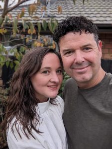 Elisabeth is a middle aged Pākahā woman, with long brown hair falling in waves over her shoulders. She is wearing a white linen blouse that is pleated into a standing collar.Ryan is a Pākehā man, also middle-aged, with black curly hair on top of his head, and shorter, greying sides. He is wearing a olive coloured tee shirt, and is sporting light stubble.Both are smiling at the camera, posed in a suburban garden, with autumn leaves turning in the background.