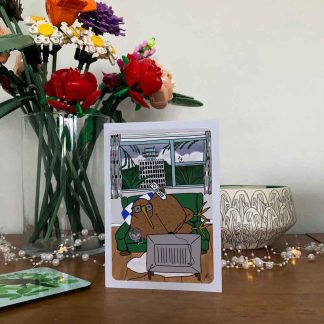 A greeting card with an illustration of a giant kiwi relaxing on the couch, remote in hand, kitty curled up under his wing. In the foreground, the back of an old-school television, in the background, the Avalon Studio Tower. The card is displayed on a wooden sideboard with various decor items.